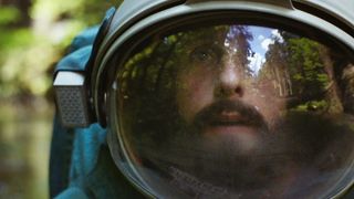 a bearded man's face can be seen through a spacesuit visor. he appears to be standing in a forest