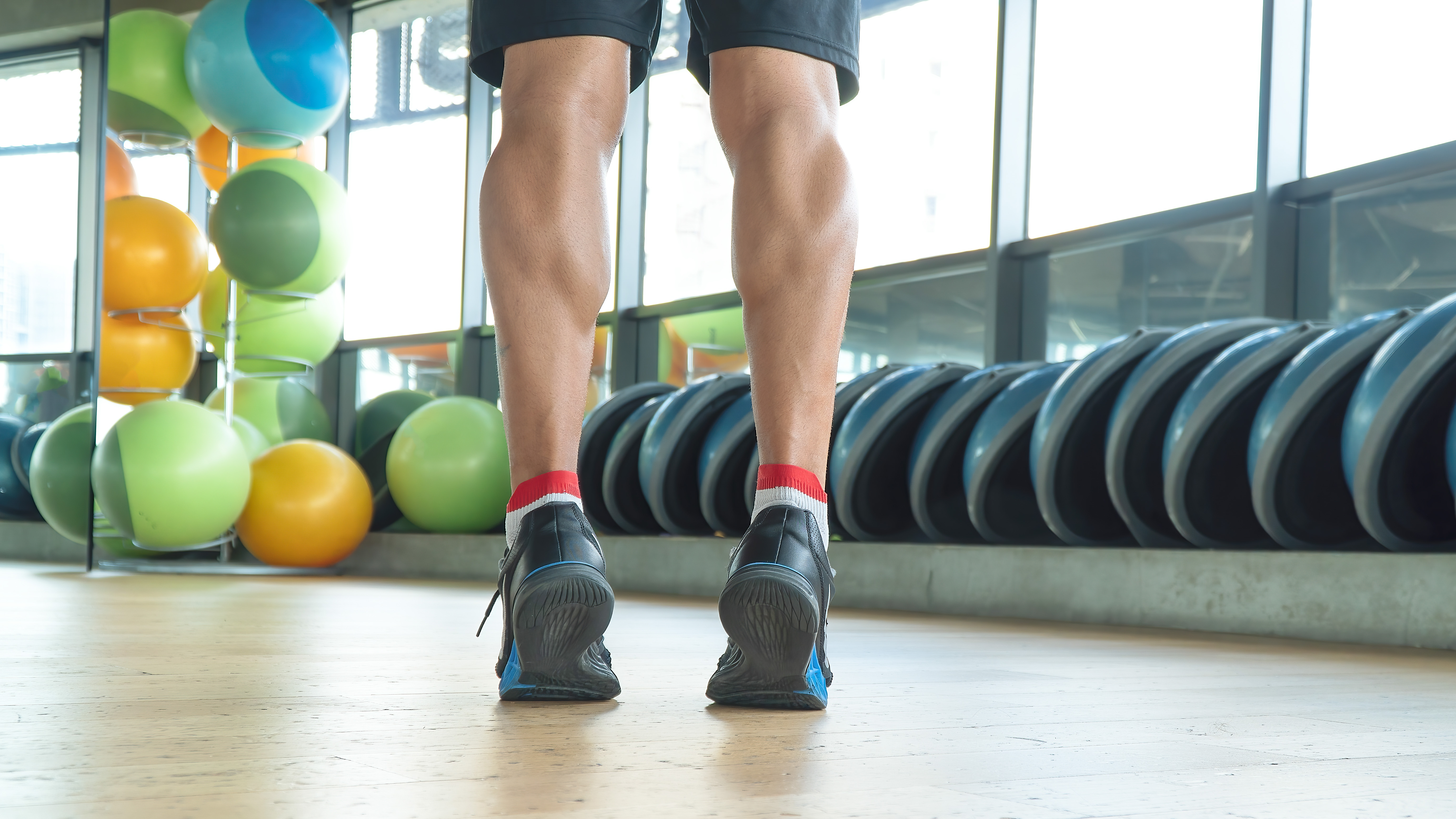 A man performs calf raises in a gym. We see the bottom of his legs from the back; he is standing on his tip toes. In front of him are balance boards and exercise balls.