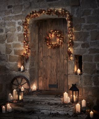 A front doorstep with candles and lanterns on the steps and a leafy garland and wreath on and around the wooden door