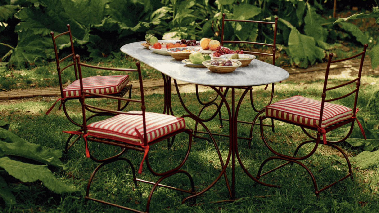 Rotating GIF of different images of charming gardens and garden decor. There is a grassy backyard with iron chairs that have red striped pillows, a close-up shot of a decorated table, and a round bistro table with glassware on it.