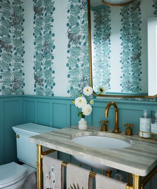 Teal bathroom with wainscotting and wallpaper, brass fixtures and fittings, marble topped vanity unit, brass mirror