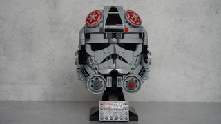 A gray helmet with black eyes and frontal features also has splashes of red on the top and the bottom is on a black display stand with a name plaque in front of a gray background