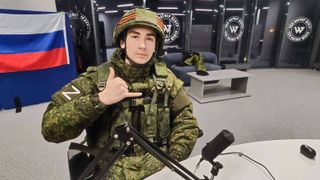 Russian streamer GrishaPutin sits in the Wagner Group headquarters in full Russian army gear, flashing a shaka sign at the camera.