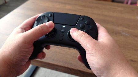 Hands holding a black gaming controller over a wooden table. The controller has visible buttons and an analog stick, suggesting gameplay action.