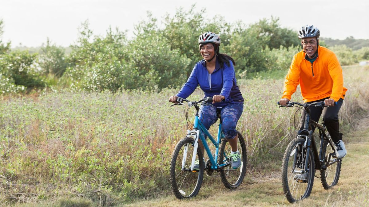 photo shows a man and woman riding bikes next to each other on a grassy path. They&#039;re both wearing helmets and smiling