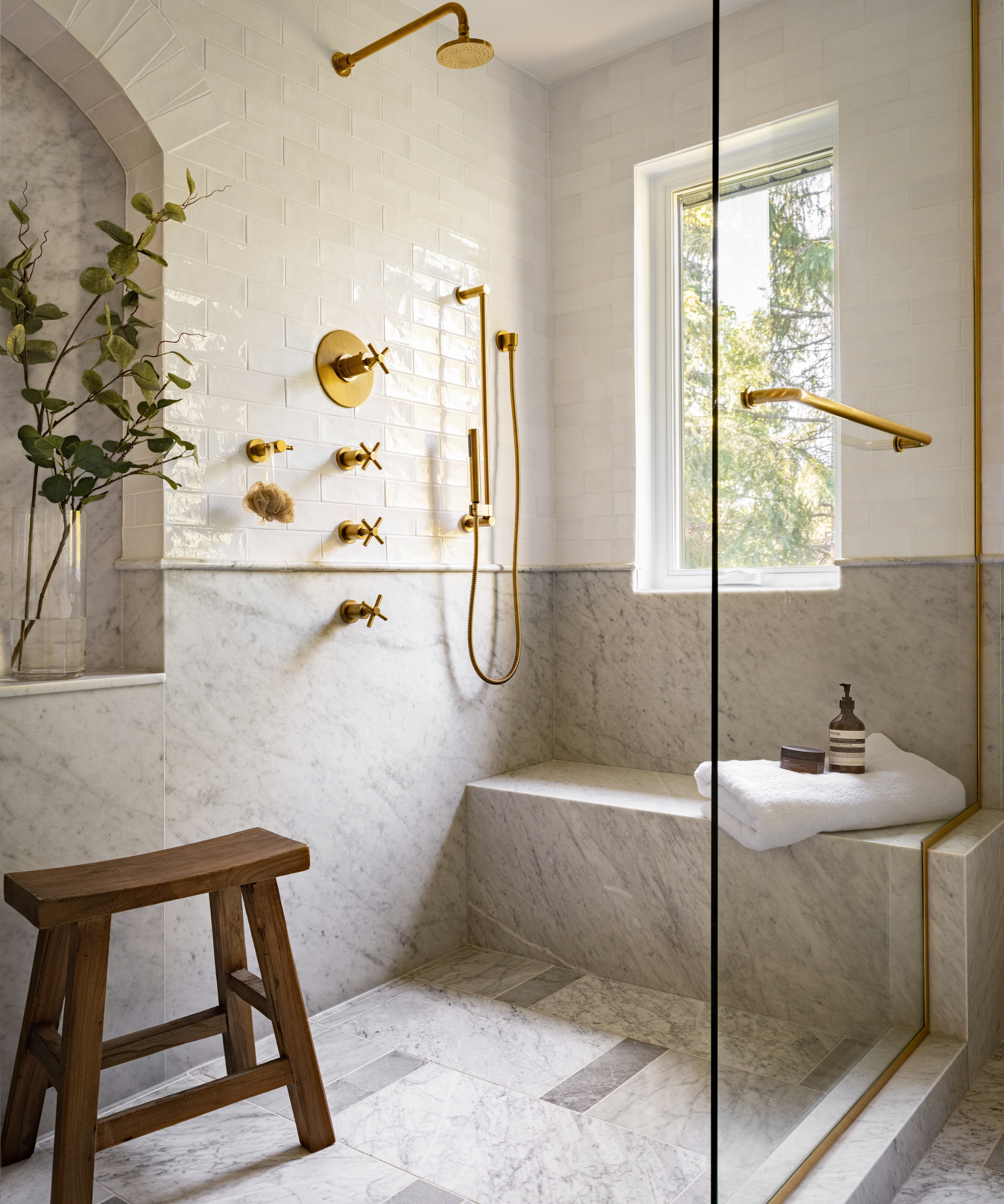 A large walk-in shower with marble floors and lower walls with an integrated shower bench. On the other side, a wooden freestanding bench sits beneath an arched shower niche