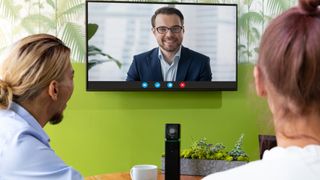 A man and woman sit in a small conference room using Boom videoconferencing solutions with a smiling man on a display.