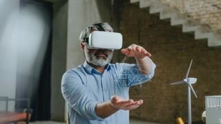 An older man who is an architect, wearing a VR headset to represent the business use cases for virtual reality. His hands are raised and a digital blueprint floats between them, while a model wind turbine is visible in the office behind him.