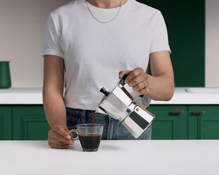 A female adult wearing a white t-shirt and gold necklace holding a moka / percolator and pouring coffee in kitchen with green cabinet decor to demonstrate how to use a coffee maker
