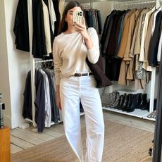 British influencer Anna Newton poses for a mirror selfie in her closet wearing a beige lightweight crewneck sweater, a brown suede tote bag, black belt, and relaxed white jeans