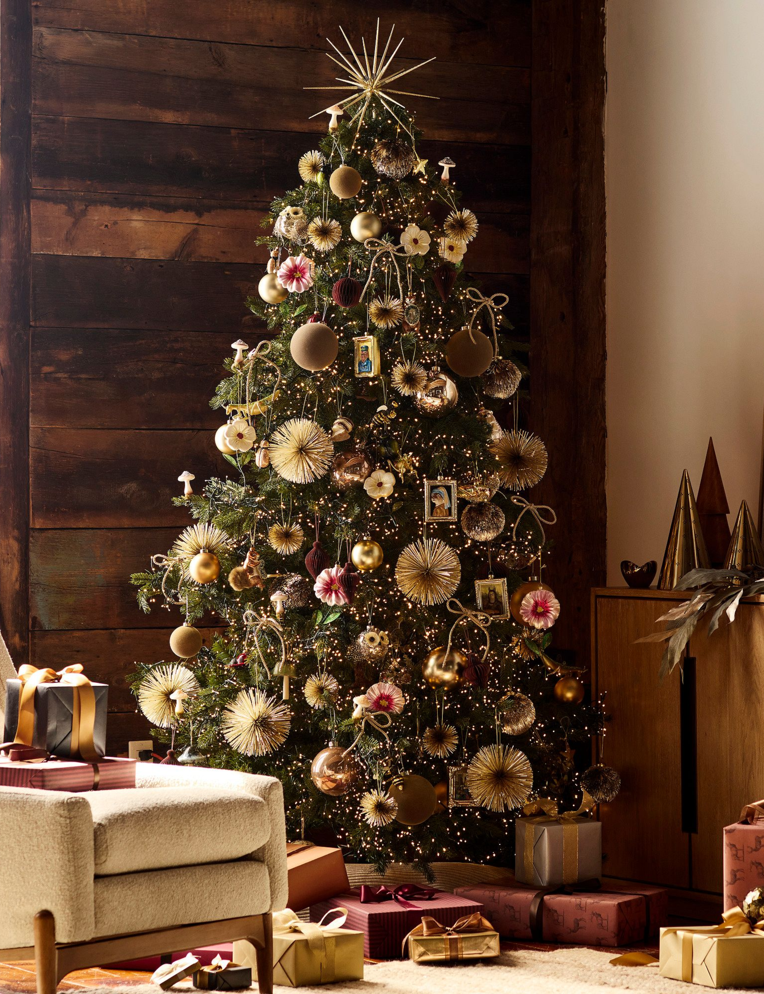 Enchanting gold-and-pink Christmas tree styled in a living room with a natural wood plank accent wall, white accent chair, and wrapped gifts.