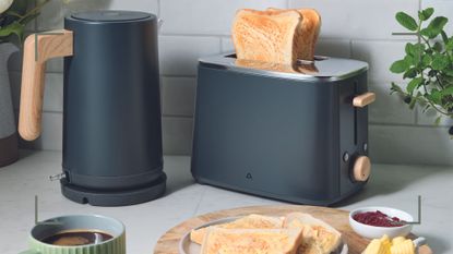 Kitchen countertop with matt grey Aldi kettle with wooden handle and matching toaster beside a wooden plate with toast to show the new Aldi kitchen appliances in stores