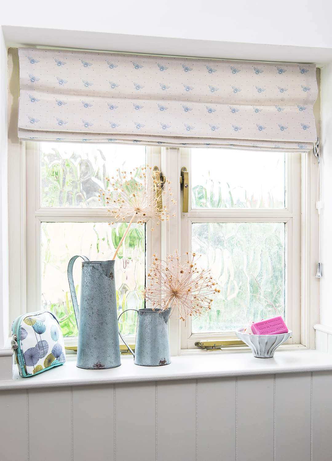 Closed window with a bee print fabric blind and two zince jugs with dried flower heads on display