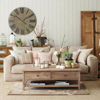 Grey panelled living room, large oversized wood clock, beige sofa, matching cushions, cream white armchair, wooden console table, rustic wood coffee table and wardrobe.