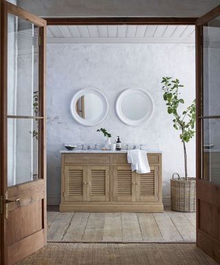 view from bedroom into ensuite bathroom. Large wooden and glass doors, natural wooden flooring, wooden twin basin with toiletries and small vase, two rounded white mirrors wall-mounted above sinks, large planter with plant to the right of the sinks, textured white painted walls.