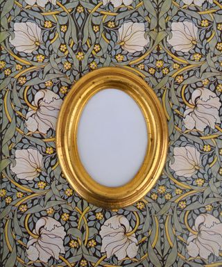 An oval shaped golden frame on top of a floral fabric with white flowers, golden vines, pale green leaves, and golden smaller flowers