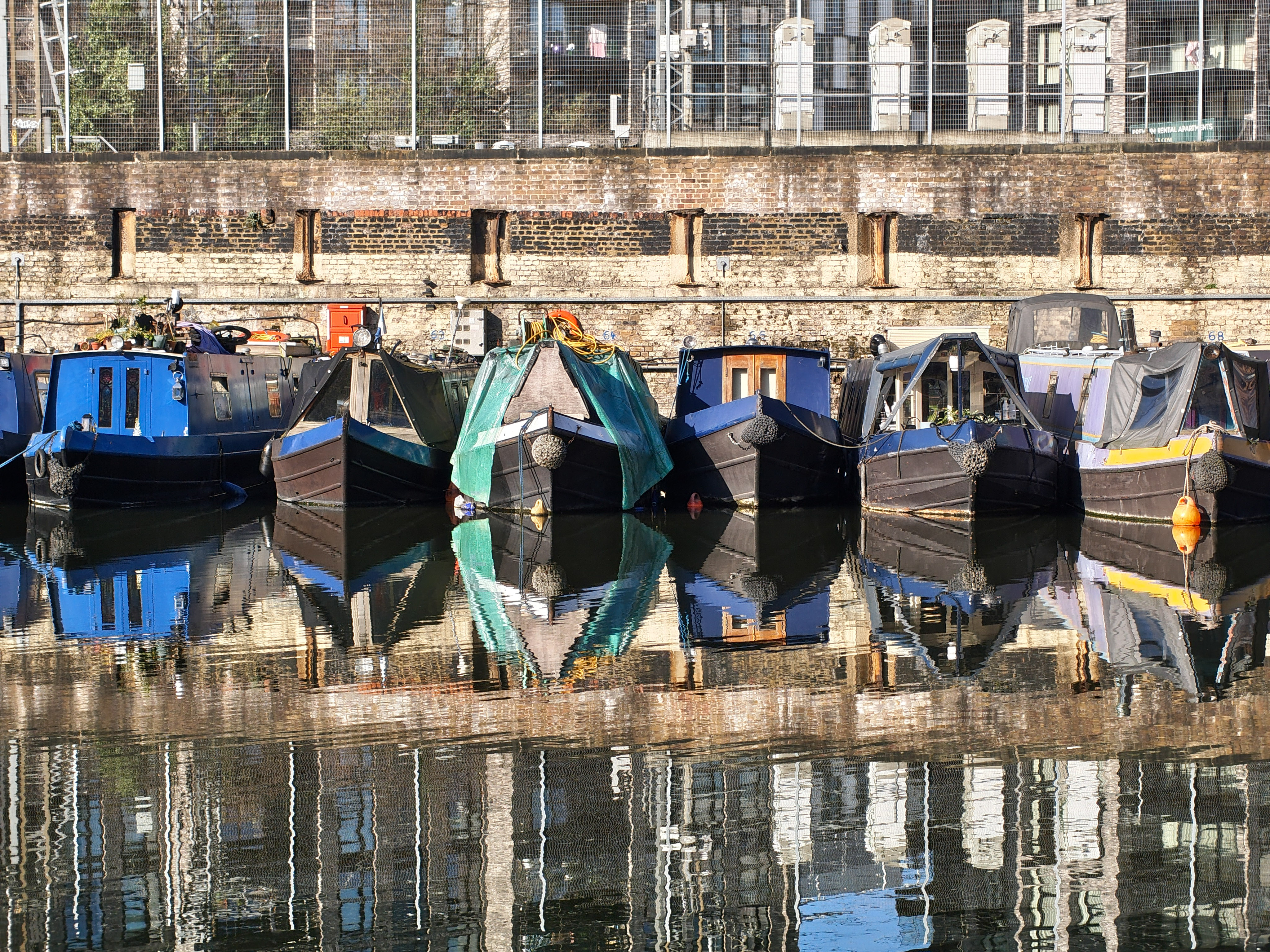Row of canal narrowboats moored side-by-side with reflections mirrored in still water, photographed with the Nothing Phone (4a).