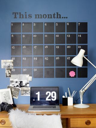 Chalkboard wall calendar above a desk with laptop and lamp in a home office, on a blue wall