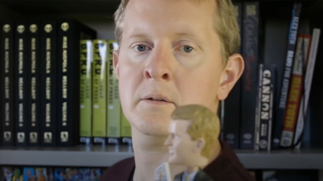A close up of Ken Jennings in front of a bookcase, with a Ken Jennings bobblehead in front of him.