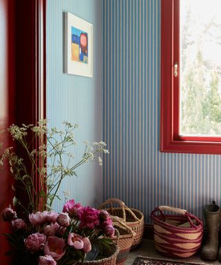 boot room with blue and white striped walls, red trim around window and woven baskets on the floor with bunches of pink flowers in the foreground