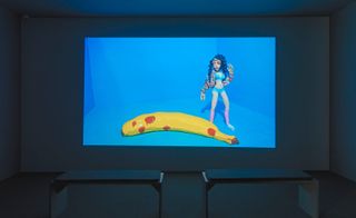 White darkened room, grey floor, two stool seats centre, projection screen on the wall of a caricature female in a bikini, jacket, socks and shoes, long wavy black hair, model banana on the ground against a blue background