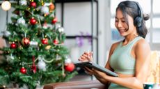A young woman dressed in activewear is sitting next to a Christmas tree with headphones on and whilst holding a tablet
