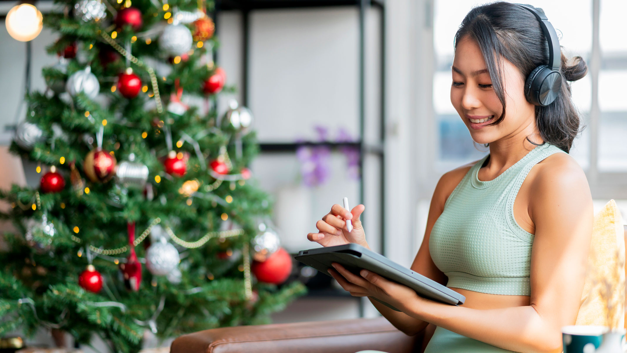 A young woman dressed in activewear is sitting next to a Christmas tree with headphones on and whilst holding a tablet