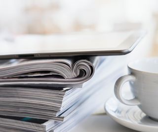 pile of magazines with electronic tablet on top and white cup and saucer to side
