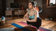 A woman performs a deep hip rocker stretch in a home setting. Her hair is tied back and she wears a teal tank-top and black yoga pants. Her feet are bare and she is balanced on a multicolored yoga mat