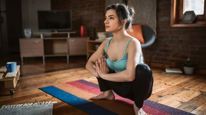 A woman performs a deep hip rocker stretch in a home setting. Her hair is tied back and she wears a teal tank-top and black yoga pants. Her feet are bare and she is balanced on a multicolored yoga mat