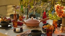 An outdoor table set with a striped tablecloth, vases of flowers, bottles of wine and amber glasses, plates of fruits and berries near a stack of set dining plates, and a terracotta-colored pan on a heat-safe holder in the middle