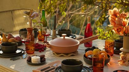 An outdoor table set with a striped tablecloth, vases of flowers, bottles of wine and amber glasses, plates of fruits and berries near a stack of set dining plates, and a terracotta-colored pan on a heat-safe holder in the middle