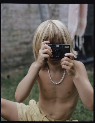 Child wears a pearl necklace and points camera