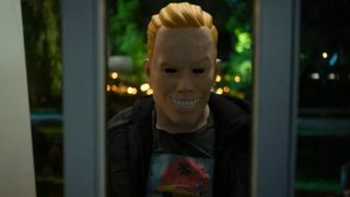 A teenager wearing a mask of a smiling man and standing outside a window during the Amazon Prime horror movie, Totally Killer.