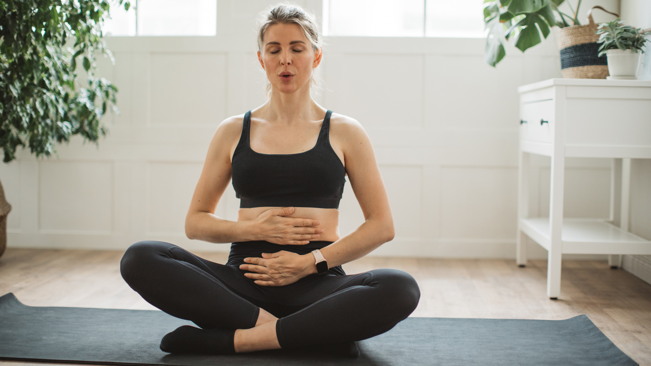 Woman sits cross legged on an exercie mat in a domestic setting with her hands on her belly and her lips pursed as she exhales