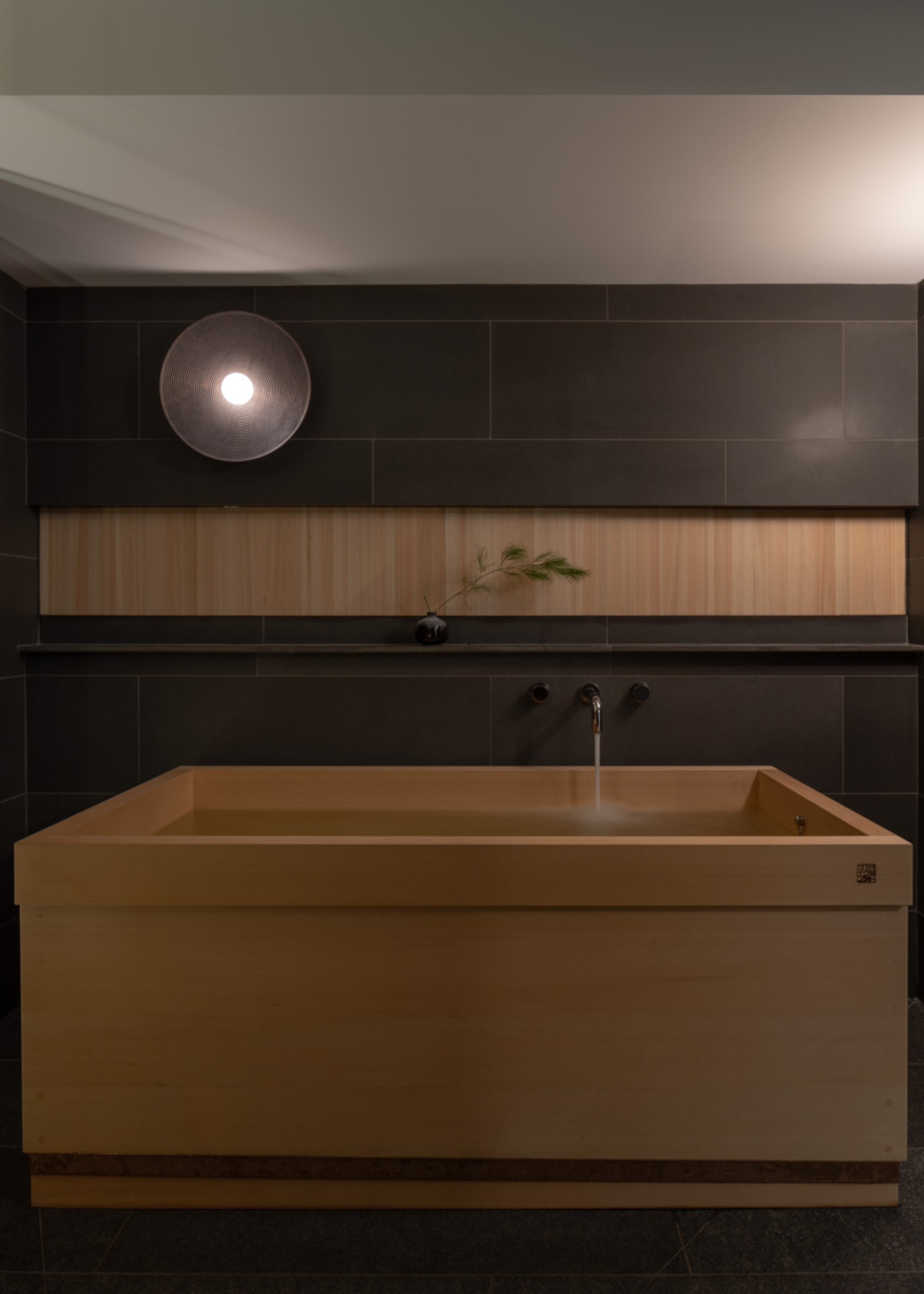 A Japanese bathtub with a flowing tap, a black tiled wall, a wood panel, and a round wall sconce