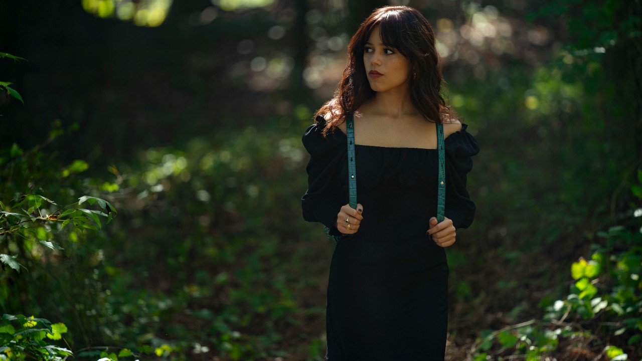 A press photo of Jenna Ortega walking through the woods holding the straps of her backpack in Miller's Girl.