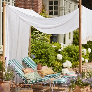 Canopy created with white tasseled fabric strung between four wooden upright posts, above two sun loungers with green and white striped cushions, and hydrangeas in the background