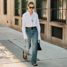street style NYFW SS26 of fashion editor walking wearing black sunglasses, a white button-down shirt, a cord necklace with silver pendant, black belt, jeans, a black bag, and black flip-flop sandals