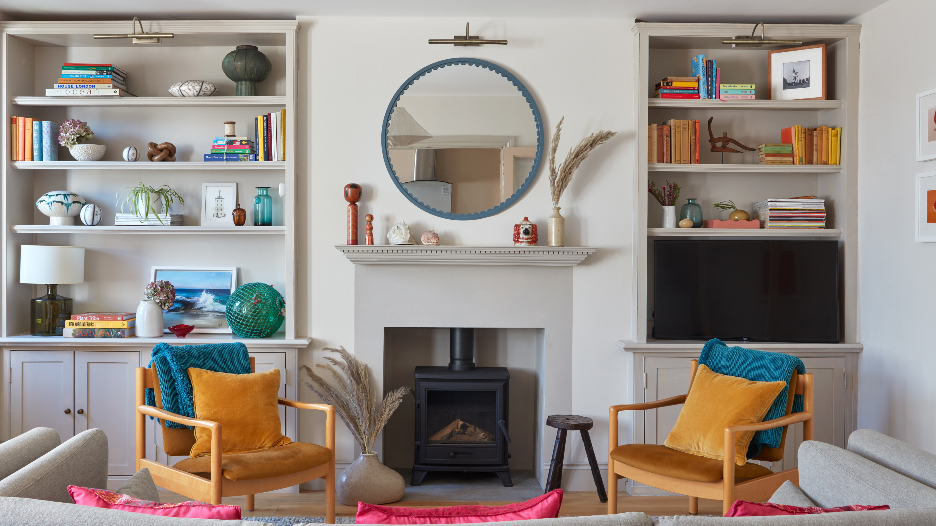 a neutral living room with pops of bright colour, a cream sofa, yellow mid century style armchairs and a round mirror over a fire surround and alcove bookcases either side of the chimney breast