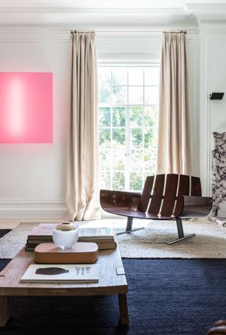 Blue and white living room with a neon pink painting hanging beside the window on the back wall. There is an oversized wooden chair in front of the window and a wooden coffee table.