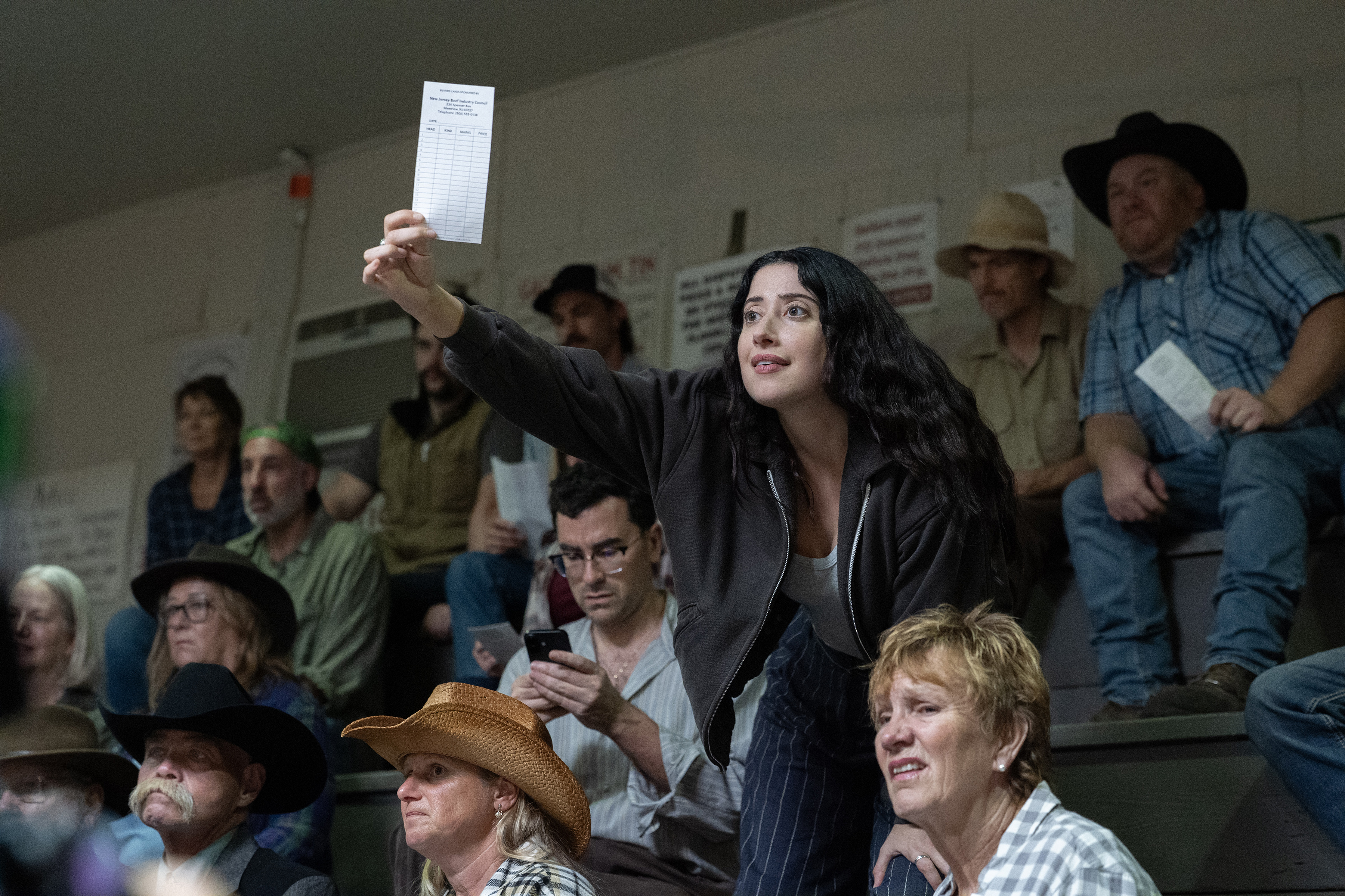 A man (Dan Levy as Nicky) looks down at his phone and a woman (Taylor Ortega as Morgan) stands while holding up a bidding slip, as they sit in a crowd at an auction, in Episode 105 of BIG MISTAKES.