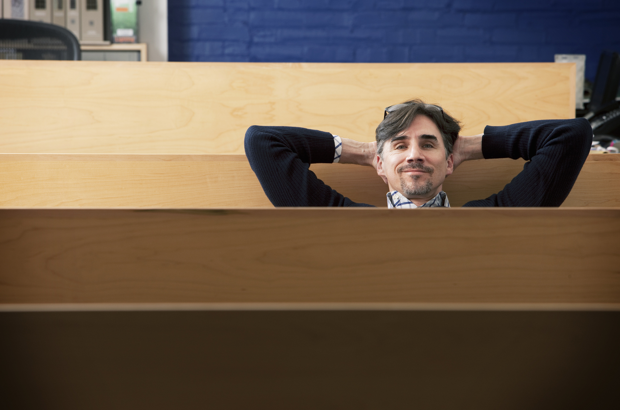A man in his 40s leans back with his hands behind his head, smiling, in an architectural services office.