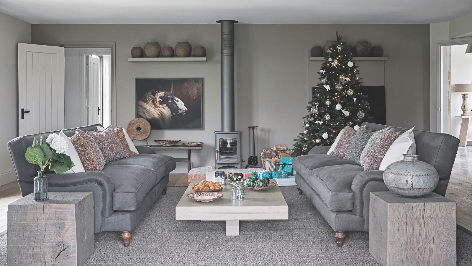 A grey living room with grey couches, walls, carpet and vases. Between the couches is a white coffee table, and behind is a wood burner. To the right of the burner is a large decorated Christmas tree, and to the left is an artwork of a ram and an open doorway.