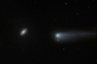 a comet on the right has a red/pink nucleus in the center and a glowing greeny blue coma which stretches out into a long tail to the right. The black eye galaxy shines bright but has a distinct dark patch covering one side of the galaxy center.