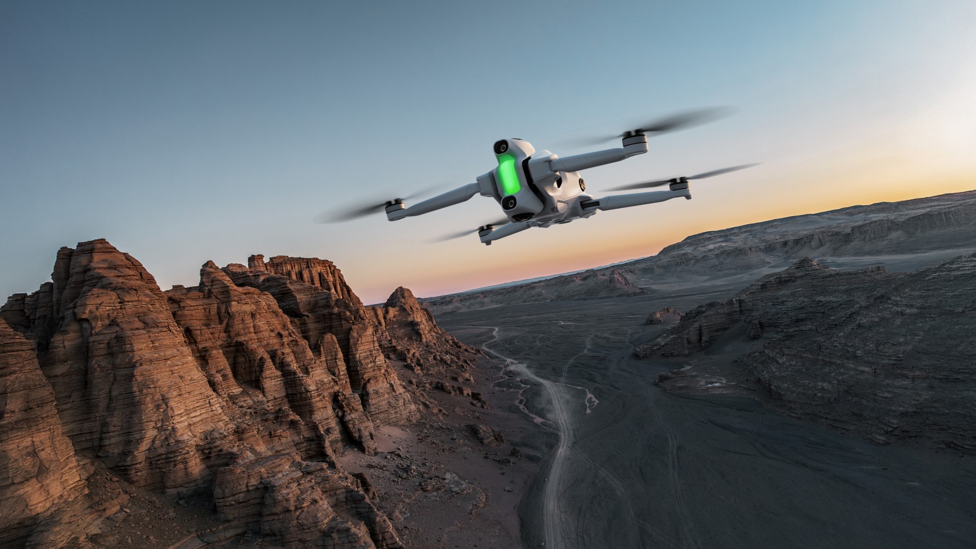 A white drone with a green light on its back flies low over a desert canyon at sunset.
