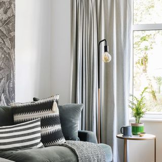 Grey velvet sofa with black and white cushions on a wooden floor with grey curtain beside window
