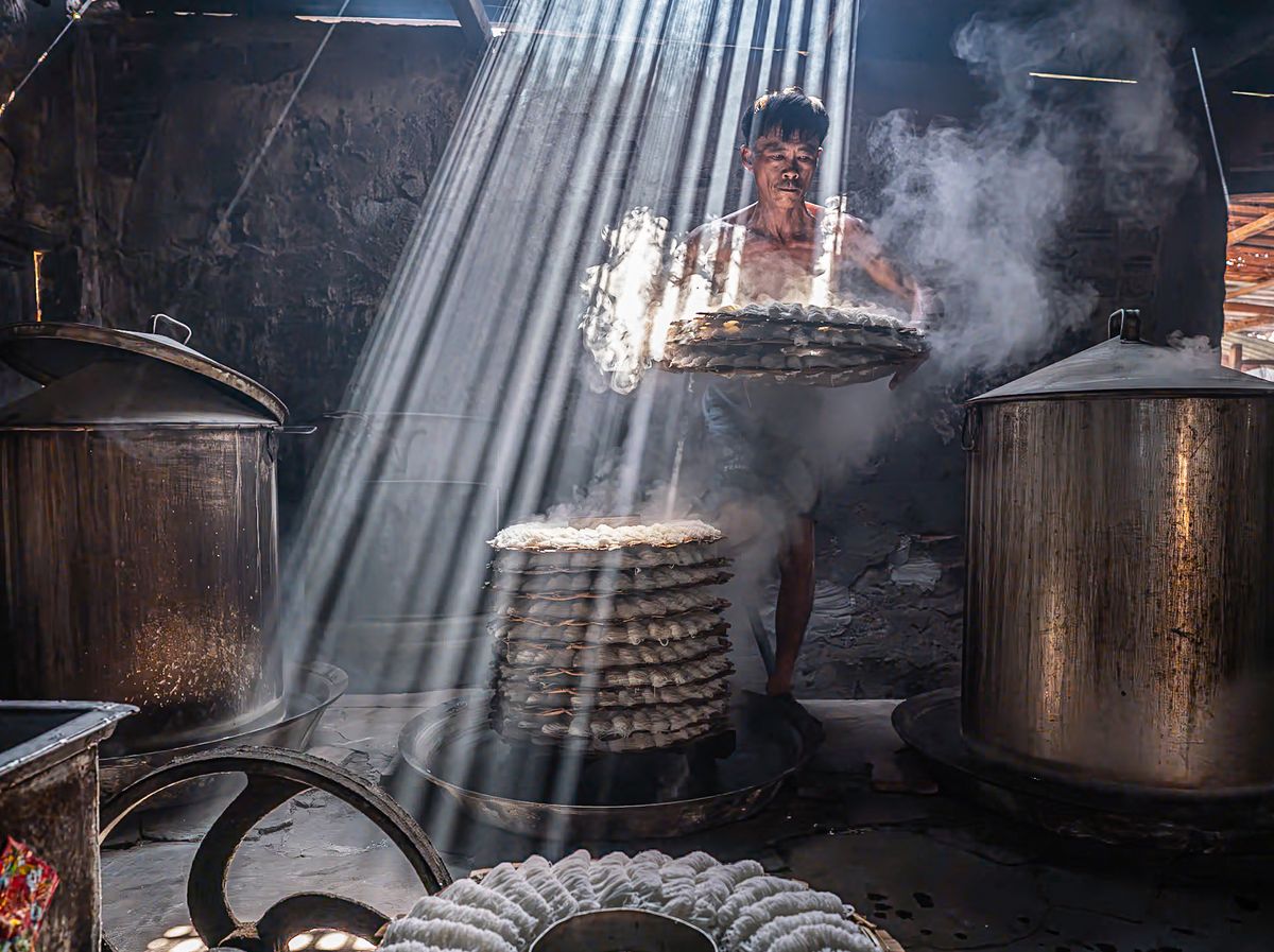A worker lifts a stack of steaming trays in a dimly lit kitchen, with rays of light illuminating the smoke-filled air around him