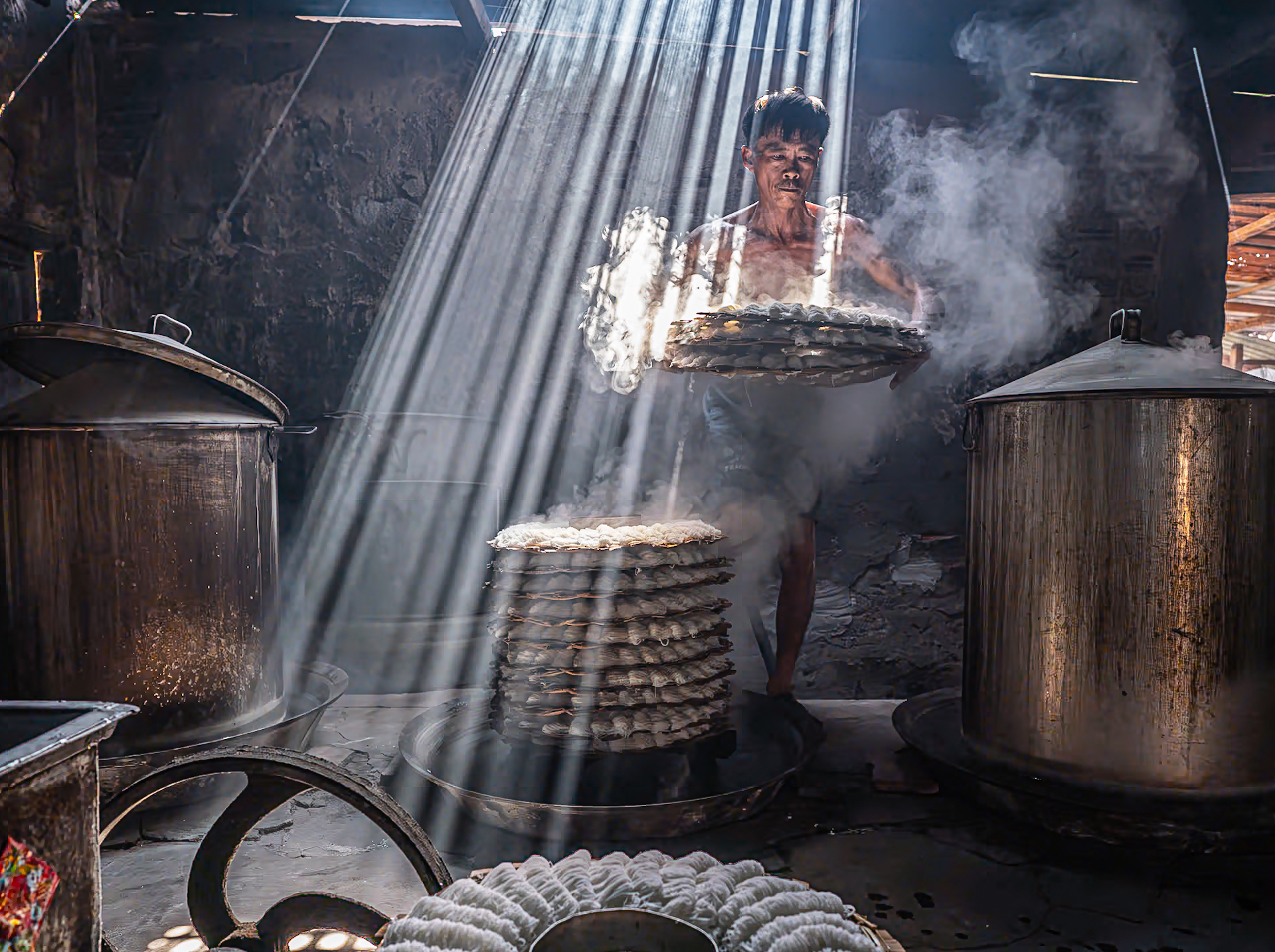 A worker lifts a stack of steaming trays in a dimly lit kitchen, with rays of light illuminating the smoke-filled air around him