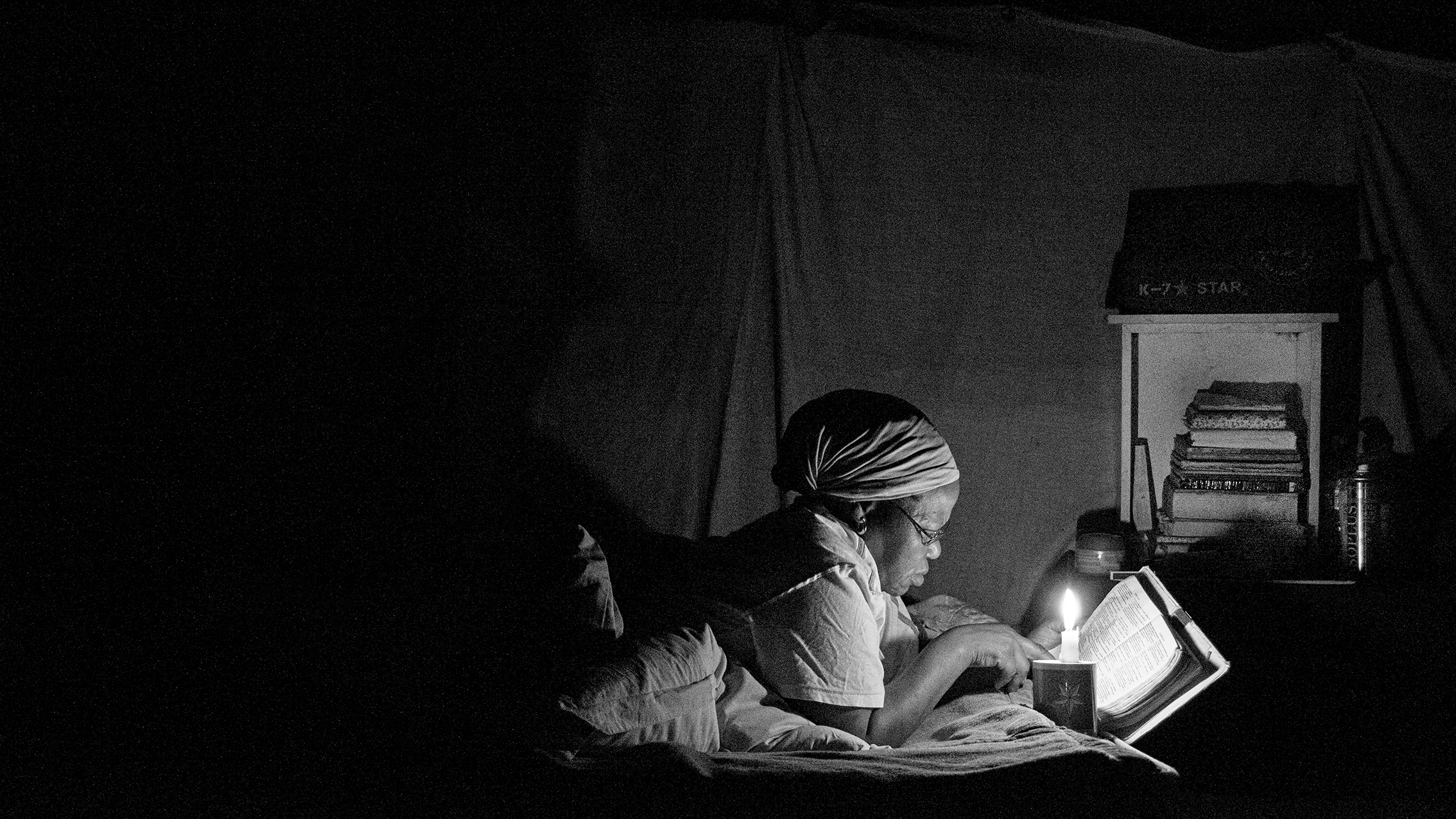 Black and white photograph of a woman lying on her stomach reading a bible by candlelight
